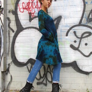 May include: A long-sleeved, tie-dye tunic in shades of blue and green, paired with light blue jeans and black lace-up boots with a chunky heel. The background features a graffiti-covered brick wall.