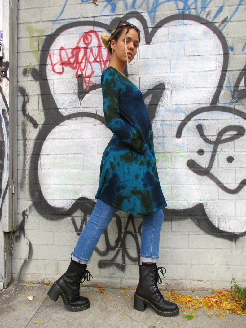 May include: A long-sleeved, tie-dye tunic in shades of blue and green, paired with light blue jeans and black lace-up boots with a chunky heel. The background features a graffiti-covered brick wall.