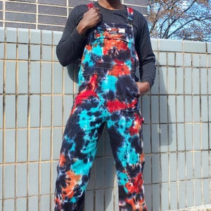 Puede incluir: Peto vaquero largo con un vibrante estampado tie-dye en tonos negro, turquesa brillante y naranja intenso. La prenda incluye tirantes ajustables, un bolsillo en el peto delantero y bolsillos laterales, mostrando una estética audaz y colorida.