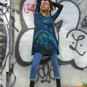 May include: A tie-dye tunic in blue and green, worn with cropped blue jeans and black boots. The model is standing in front of a brick wall covered in graffiti.