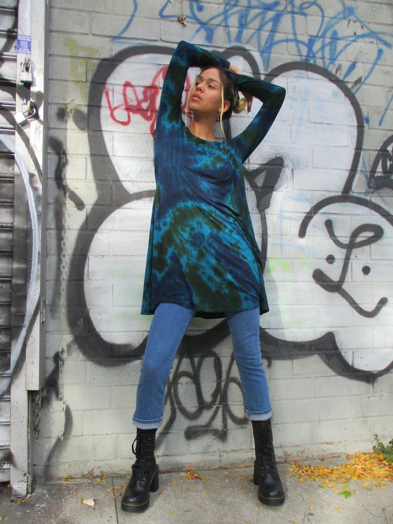May include: A tie-dye tunic in blue and green, worn with cropped blue jeans and black boots. The model is standing in front of a brick wall covered in graffiti.