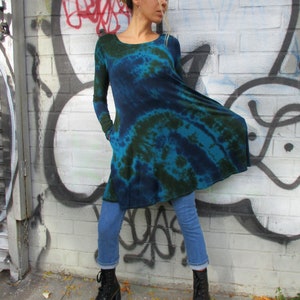 May include: A long-sleeved, tie-dye tunic in teal and navy blue. The tunic has a scoop neck and pockets. The model is wearing blue jeans and black lace-up boots. The background is a brick wall with graffiti.