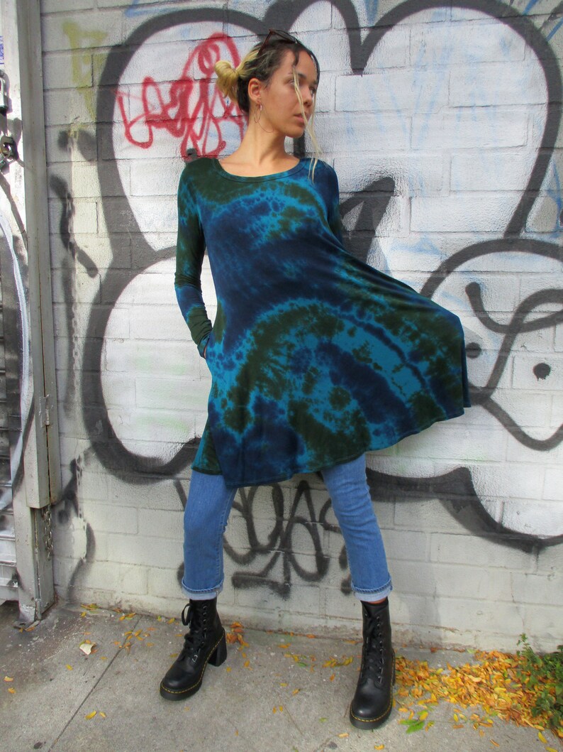 May include: A long-sleeved, tie-dye tunic in teal and navy blue. The tunic has a scoop neck and pockets. The model is wearing blue jeans and black lace-up boots. The background is a brick wall with graffiti.