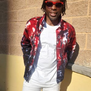 Puede incluir: Chaqueta vaquera teñida con efecto tie-dye en tonos rojos, negros y blancos, sobre una camiseta blanca y combinada con pantalones blancos. La chaqueta tiene botones en la parte delantera y cuello. La persona lleva gafas de sol con montura roja.