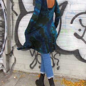 May include: A long-sleeved, tie-dye tunic in shades of blue and green. The tunic has a low back and is paired with light blue jeans and black boots. The background features a graffiti-covered brick wall.