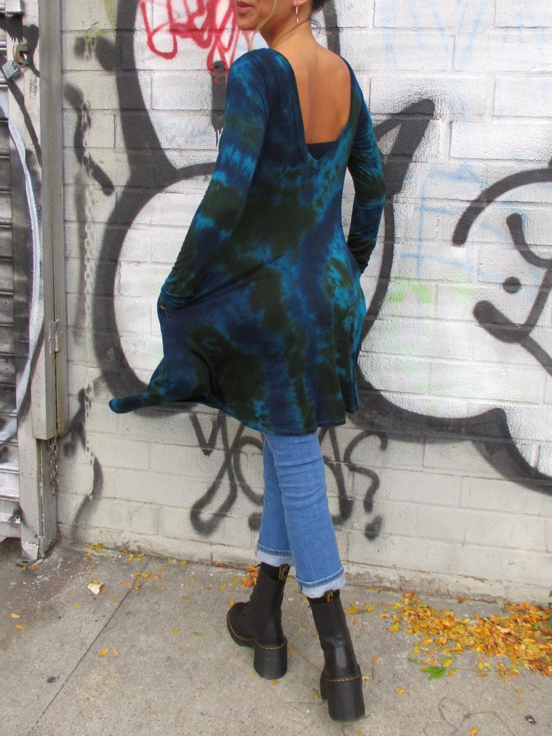 May include: A long-sleeved, tie-dye tunic in shades of blue and green. The tunic has a low back and is paired with light blue jeans and black boots. The background features a graffiti-covered brick wall.