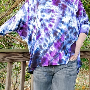 May include: A purple, blue, and white tie-dye shirt with a relaxed fit. The shirt has a round neckline and three-quarter sleeves.
