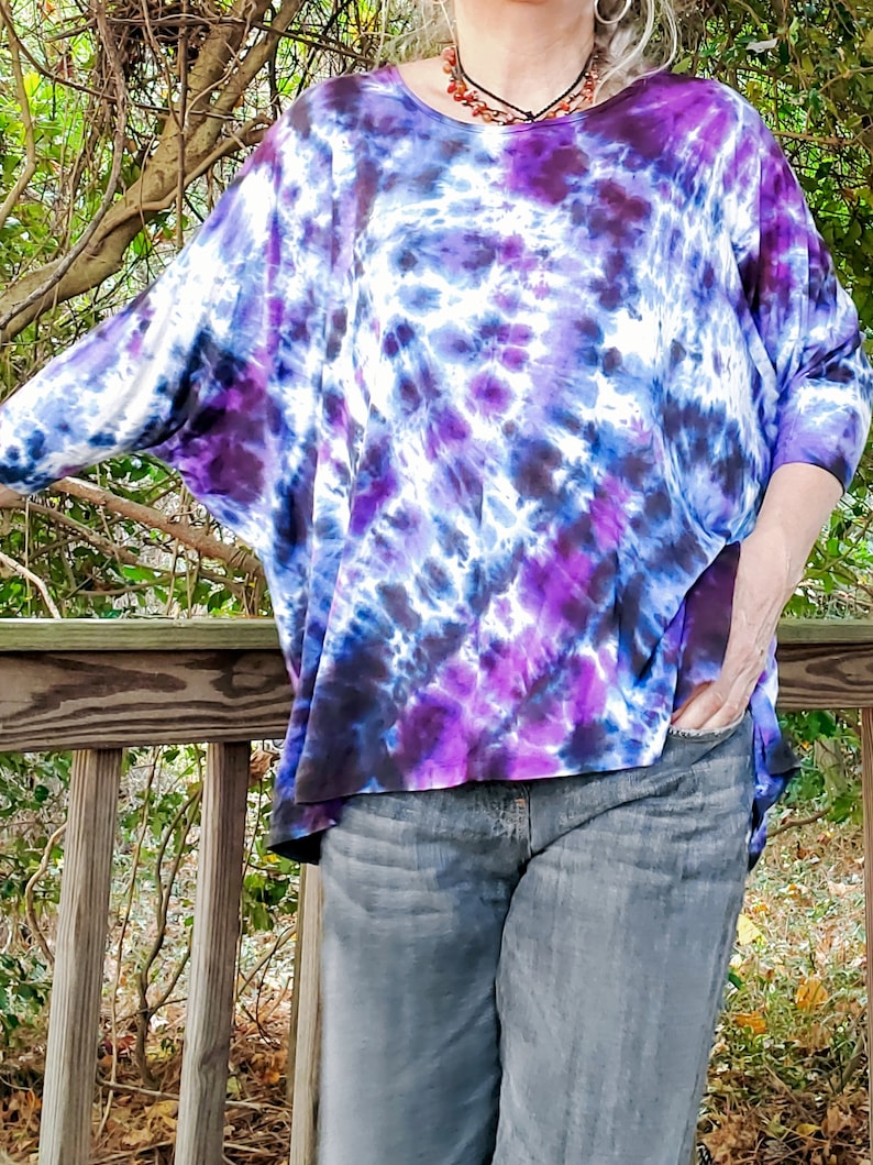 May include: A purple, blue, and white tie-dye shirt with a relaxed fit. The shirt has a round neckline and three-quarter sleeves.