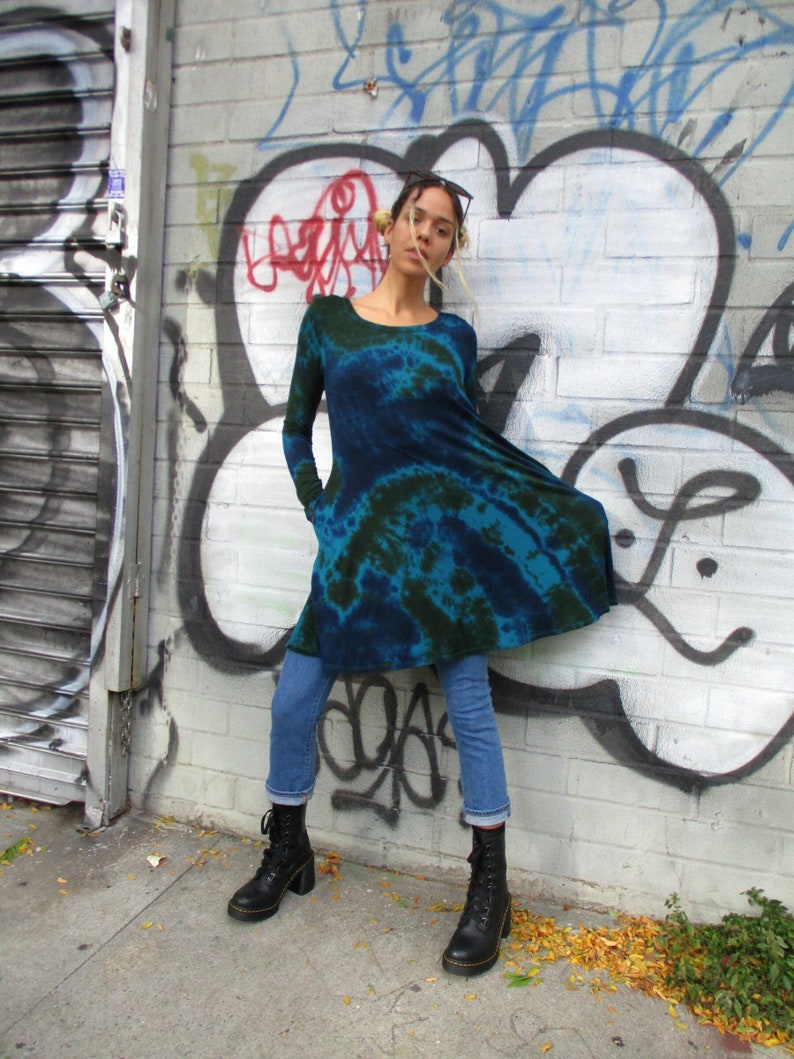 May include: A long-sleeved, tie-dye dress in shades of blue and green. The dress has a scoop neck and pockets. Worn with blue jeans and black lace-up boots. The background features a graffiti-covered wall.