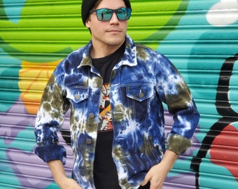 May include: A person wearing a black beanie, blue-tinted sunglasses, a blue and green tie-dye denim jacket, a black t-shirt, and black jeans. The person is standing in front of a colorful graffiti wall.