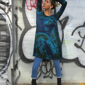 May include: A teal and dark green tie-dye tunic with long sleeves. The tunic is worn with light blue jeans and black lace-up boots. The background features a graffiti-covered brick wall.