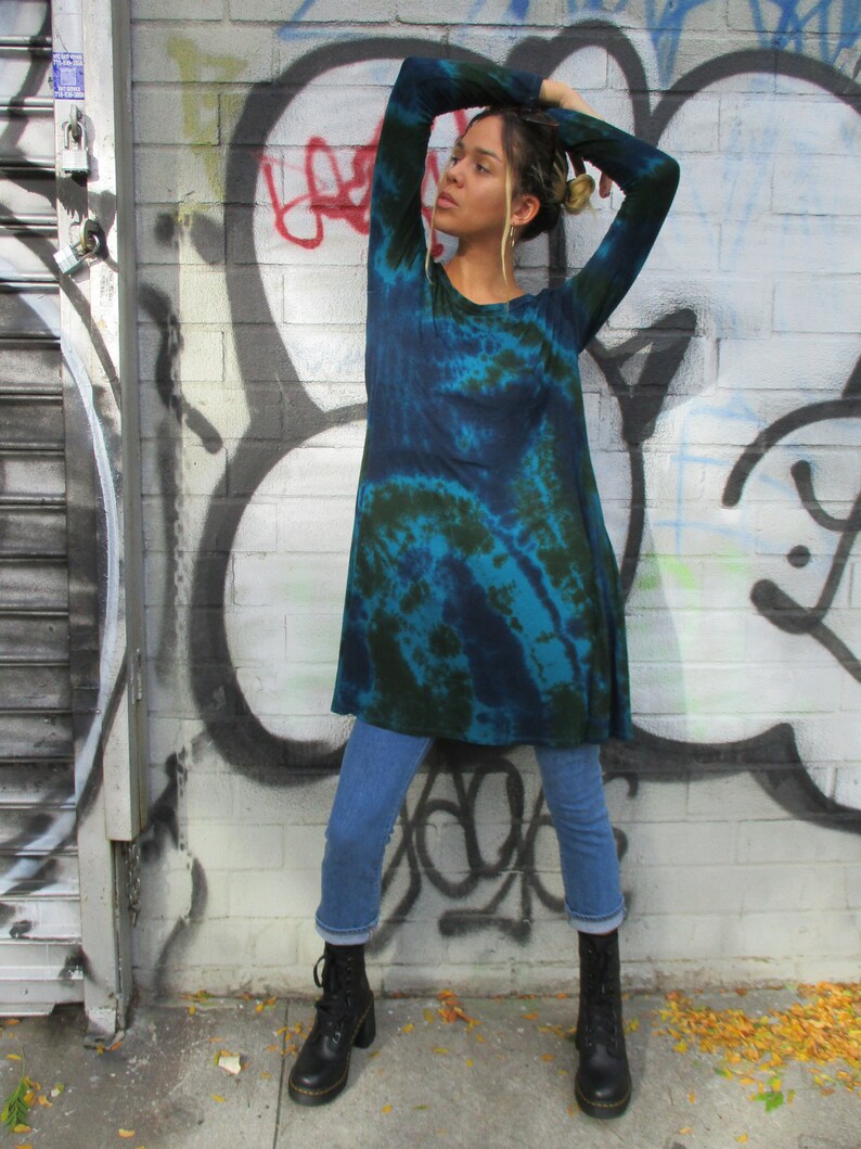 May include: A teal and dark green tie-dye tunic with long sleeves. The tunic is worn with light blue jeans and black lace-up boots. The background features a graffiti-covered brick wall.