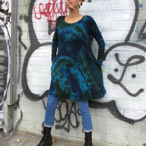 May include: A person wearing a long-sleeved, tie-dye dress in shades of blue and green, with blue jeans and black combat boots. The dress has a scoop neck and pockets. The background is a graffiti-covered wall.