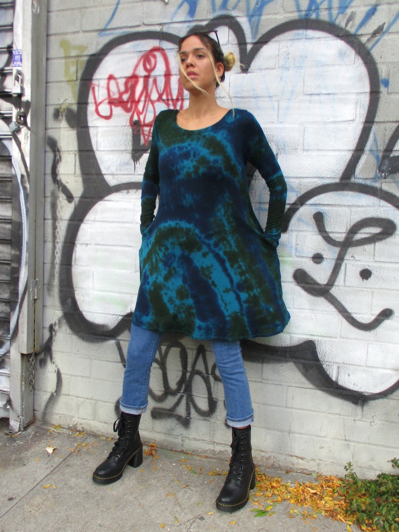 May include: A person wearing a long-sleeved, tie-dye dress in shades of blue and green, with blue jeans and black combat boots. The dress has a scoop neck and pockets. The background is a graffiti-covered wall.