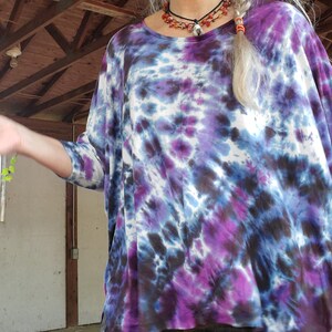 May include: A woman wearing a purple, blue, and white tie-dye shirt with a long braid. She is wearing a layered necklace with a pendant.