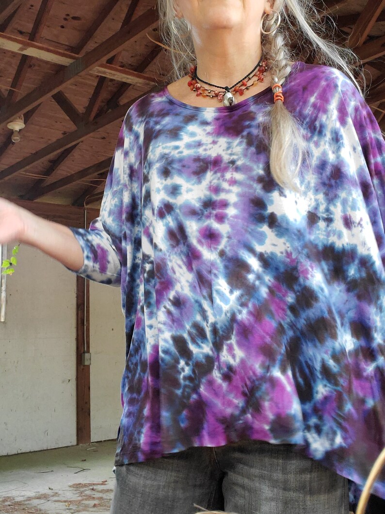 May include: A woman wearing a purple, blue, and white tie-dye shirt with a long braid. She is wearing a layered necklace with a pendant.