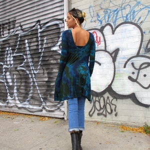 May include: A blue and green tie-dye tunic with long sleeves, worn with light blue jeans and black boots. The tunic features a low scoop back and an uneven hem. The background shows a wall covered in graffiti, with the model walking on a pavement.