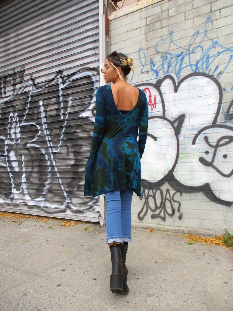 May include: A blue and green tie-dye tunic with long sleeves, worn with light blue jeans and black boots. The tunic features a low scoop back and an uneven hem. The background shows a wall covered in graffiti, with the model walking on a pavement.
