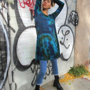 May include: A blue and green tie-dye tunic. The long-sleeved tunic is worn with blue jeans and black boots. The background is a brick wall covered in graffiti.
