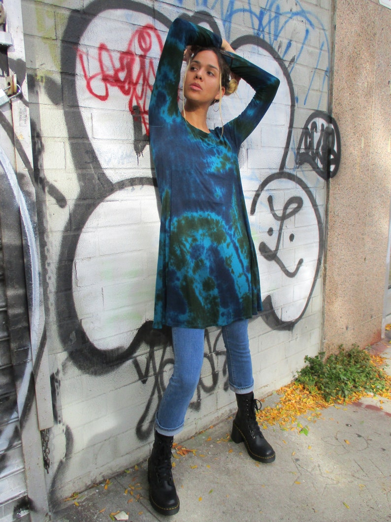May include: A blue and green tie-dye tunic. The long-sleeved tunic is worn with blue jeans and black boots. The background is a brick wall covered in graffiti.