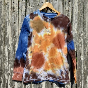 May include: A long-sleeved Henley shirt with a tie-dye pattern in shades of blue, brown, and orange. The shirt is hanging on a wooden hanger against a weathered wooden fence. The shirt has a button-up neckline.