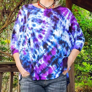 May include: A woman wearing a purple, blue, and white tie-dye shirt with a black and white pattern. The shirt has a loose fit and is made of a soft, comfortable fabric. The woman is wearing blue jeans and has her hands in her pockets.