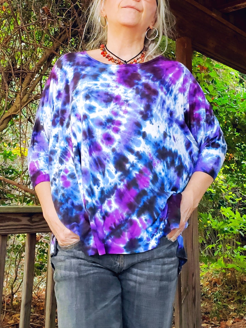 May include: A woman wearing a purple, blue, and white tie-dye shirt with a black and white pattern. The shirt has a loose fit and is made of a soft, comfortable fabric. The woman is wearing blue jeans and has her hands in her pockets.