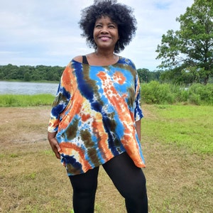 May include: A woman wearing a blue, orange, and green tie-dye shirt with black leggings. She is standing in a grassy field with a lake in the background.