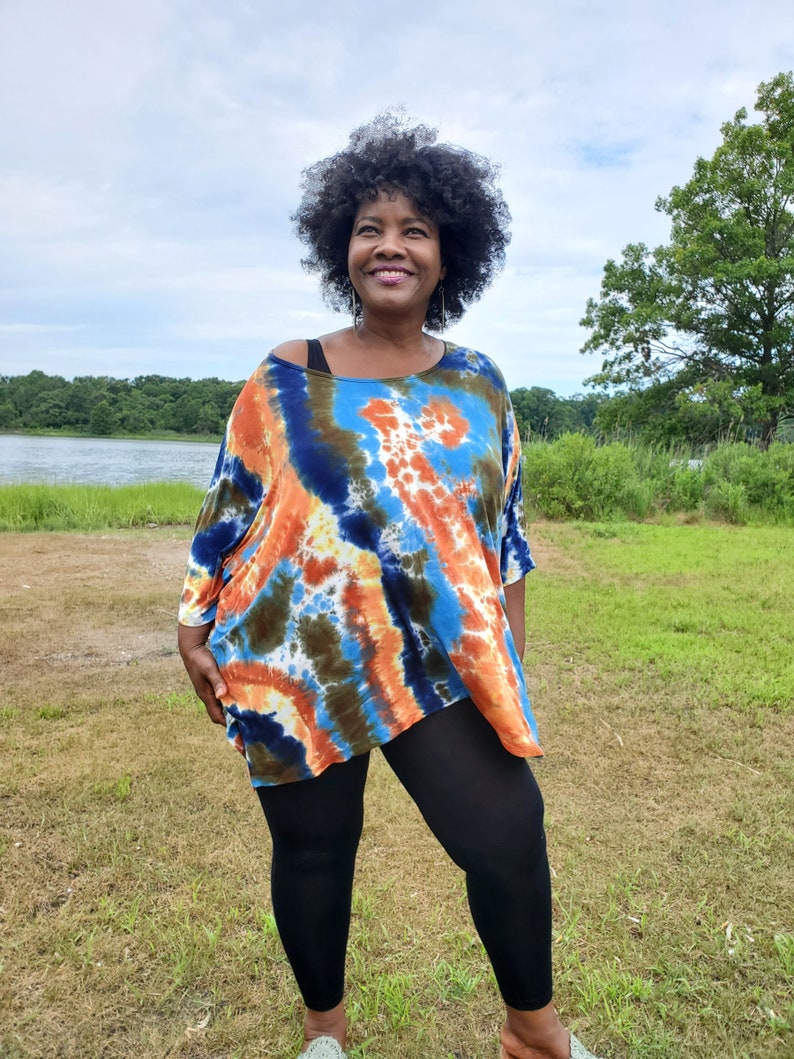 May include: A woman wearing a blue, orange, and green tie-dye shirt with black leggings. She is standing in a grassy field with a lake in the background.