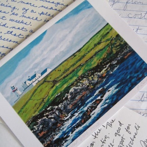 May include: A landscape art print featuring a coastal scene with a lighthouse, green fields, and blue ocean. The artwork depicts a white lighthouse and buildings on a green hillside, with a rocky shoreline and blue water.