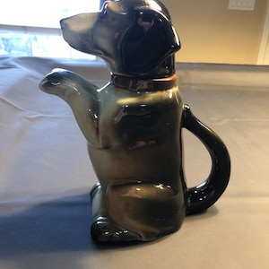 Puede incluir: Una tetera de cerámica con forma de perro y acabado brillante. La tetera es principalmente marrón oscuro y negra, con un collar marrón claro. El pico está a la izquierda y el asa a la derecha. El perro está sentado.