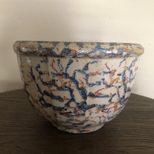 May include: A ceramic bowl with a white base and a pattern of blue and brown splatters. The bowl has a rounded shape with a slightly flared rim. The bowl is sitting on a dark wooden surface.