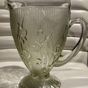May include: A clear glass pitcher featuring a raised relief pattern of iris flowers and leaves. The body of the pitcher has vertical ribbed detailing, a wide pouring spout, a sturdy handle, and a circular pedestal base with a fluted design.