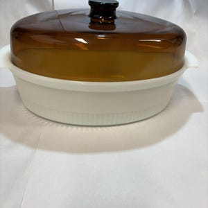 Puede incluir: Una cacerola de cerámica blanca con una tapa de vidrio marrón. La tapa tiene un pomo negro.