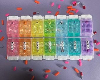 May include: A multi-coloured weekly pill organiser with compartments labelled for each day of the week. Each compartment is decorated with rhinestones and a symbol. Scattered pills in various colours surround the organiser.