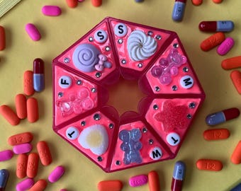 Op de afbeelding: Een roze pillenorganizer met meerdere compartimenten, versierd met diverse versieringen, omringd door oranje, roze en blauwe pillen. De organizer heeft een centraal ontwerp met een slak, een spiraal en andere vormen.