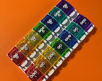 May include: A rectangular pill organiser with fourteen individual compartments arranged in two rows. Each compartment is a different colour of the rainbow, featuring white circular labels with black text indicating days of the week and AM or PM.