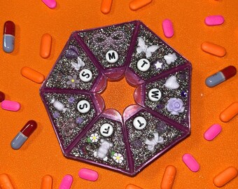Op de afbeelding: Een paarse, glinsterende pillenorganizer met zeven vakken, elk gemarkeerd met een dag van de week. De organizer is omgeven door oranje, roze en blauwe pillen. Decoratieve elementen zijn vlinders en bloemen.