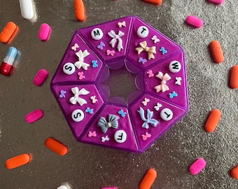 Op de afbeelding: Een paarse pillenorganizer met zeven compartimenten, versierd met strikken en de dagen van de week. Verschillende pillen in roze, oranje, wit en een blauw-rode capsule liggen verspreid rond de organizer op een zilveren oppervlak.