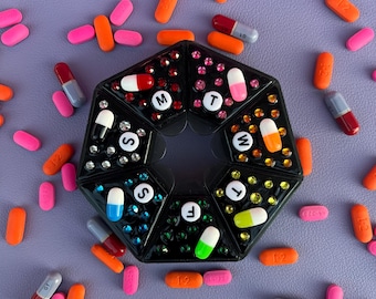 May include: A black, octagonal pill organiser with compartments labelled for each day of the week. The organiser is decorated with rhinestones and surrounded by various colourful pills and capsules in shades of pink, orange, and grey on a purple surface.