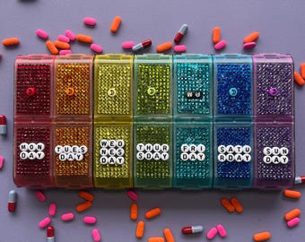 May include: A rainbow-coloured pill organiser with two rows of seven compartments, each covered in matching rhinestones. The bottom row features white letter beads spelling out the days of the week, while the top row has small moon icons.