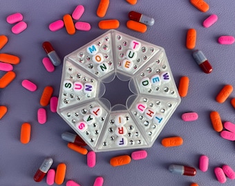 Op de afbeelding: Een heldere, achthoekige pillenorganizer met de dagen van de week in kleurrijke kralen. Verschillende roze, oranje en rode pillen en capsules liggen verspreid rond de organizer op een paars oppervlak.