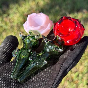 May include: Two glass pipes shaped like roses, one pink and one red, with green stems and leaves. The pipes are held in a black hand.