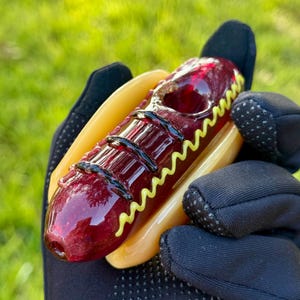May include: A glass pipe shaped like a hot dog with a red glass hot dog and a yellow glass mustard topping. The pipe is held in a black glove.