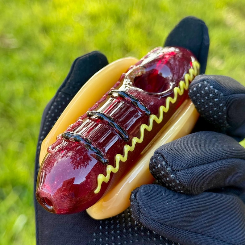May include: A glass pipe shaped like a hot dog with a red glass hot dog and a yellow glass mustard topping. The pipe is held in a black glove.