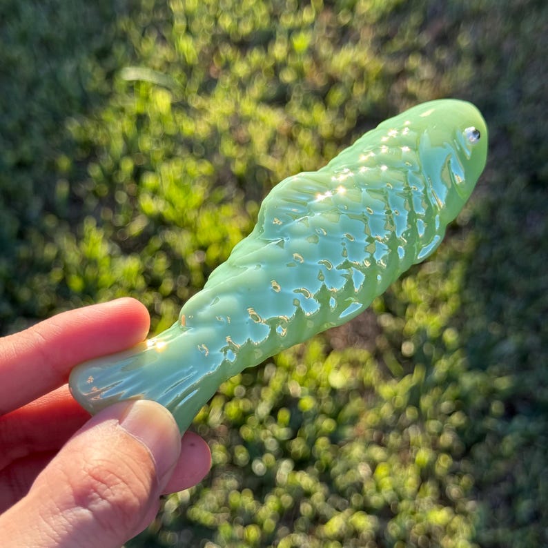 May include: A light green glass fish figurine with a textured surface. The fish has a small, round eye and a curved tail. The figurine is held in a hand against a blurred green background.