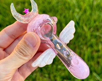 May include: A light pink glass pipe featuring a crescent moon top with a small magenta star, two white glittery wings on the sides, and a clear circular bowl. The glass has a translucent finish with subtle sparkles throughout the structure.