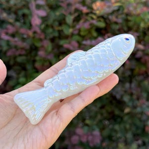 May include: A pearlescent white fish-shaped decorative object with iridescent highlights. The fish has detailed scales and a small blue eye. The object is held in a hand, with a blurred green and purple background.