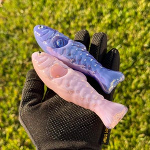 May include: Two glass fish-shaped pipes, one lavender and one blue, with silver accents. The pipes are held against a backdrop of green grass. The fish pipes have a smooth, glossy finish and are designed with intricate details.