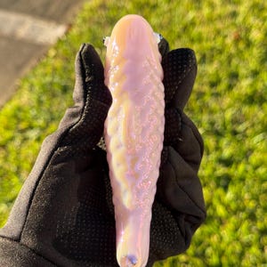 May include: A pink and white glass pipe with a textured surface. The pipe has a rounded top and a flared base. The pipe is held by a person wearing black gloves. The background is blurred green grass.